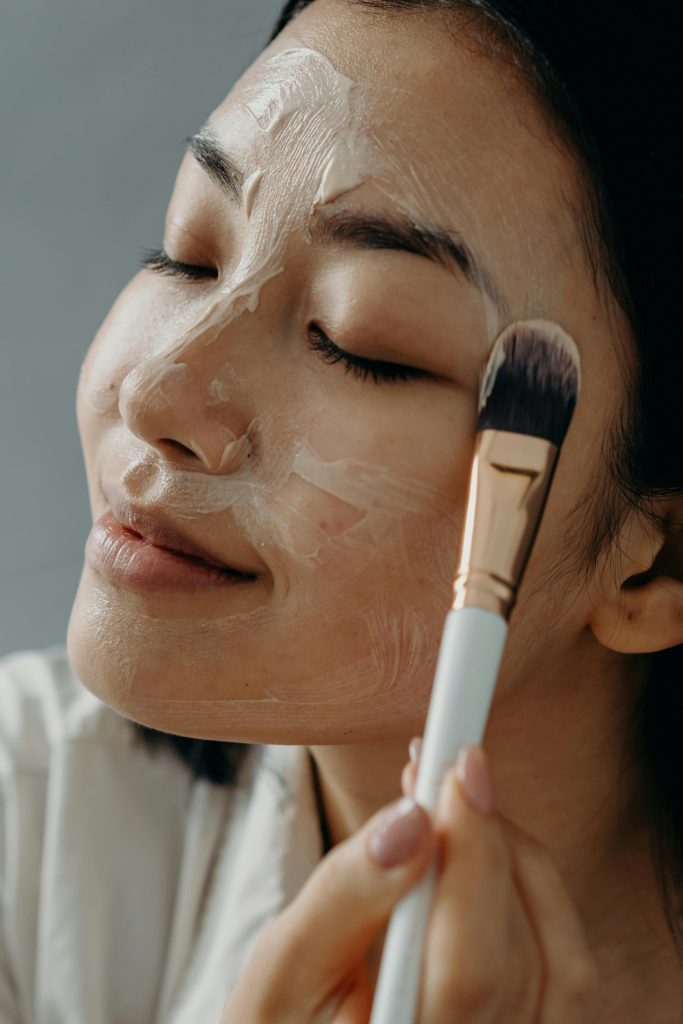 Close-up of an Asian woman enjoying a skincare routine, applying facial cream with a makeup brush for healthy skin.